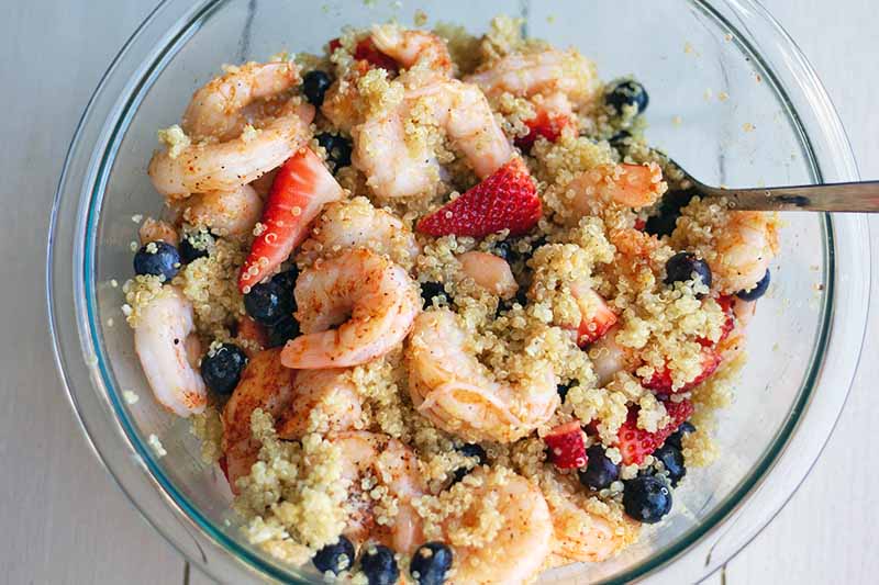 Overhead closely cropped horizontal image of a large glass mixing bowl filled with a combination of cooked quinoa and shrimp, fresh red and blue berries, and seasonings, being stirred with a metal spoon, on a white surface.