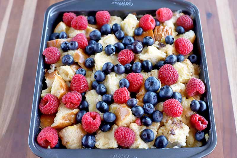 Horizontal image of a metal baking pan of berries and bread, on a striped beige and brown wood surface.
