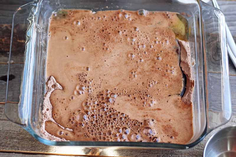 Overhead image of a frothy chocolate batter mixture in a square glass baking dish, on an unfinished wood surface.