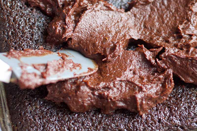 Closeup horizontal image of a spatula spreading chocolate frosting onto a cake.