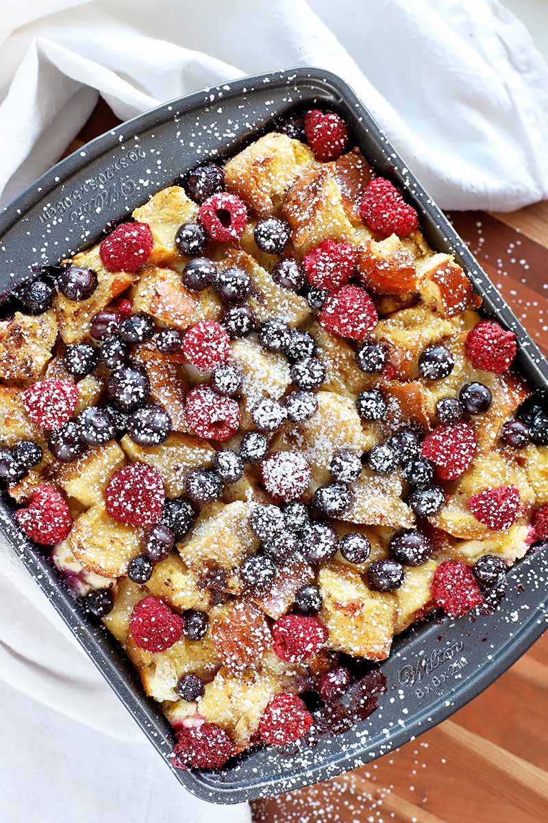 Overhead horizontal image of a metal baking pan filled with a baked breakfast casserole with fresh raspberries and blueberries, with confectioner's sugar sprinkled on top, on a wood wood surface that is partially covered with a white cloth.