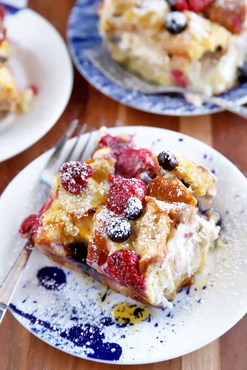Vertical image of a slice of berry breakfast casserole on a small plate with a fork in the foreground with two more similar plates in the background, on a striped wood table.