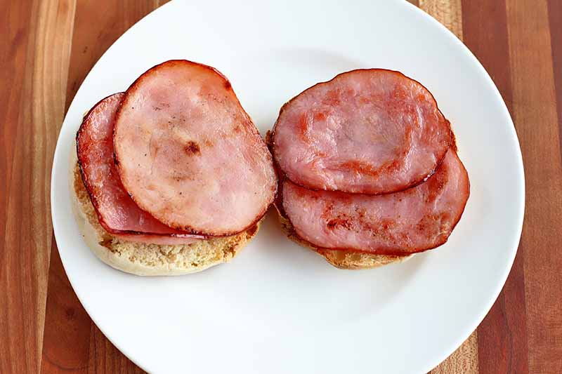 Two halves of toasted English muffin are on a white plate, topped with two slices each of Canadian ham that has been-fried until golden brown, on a wood surface.