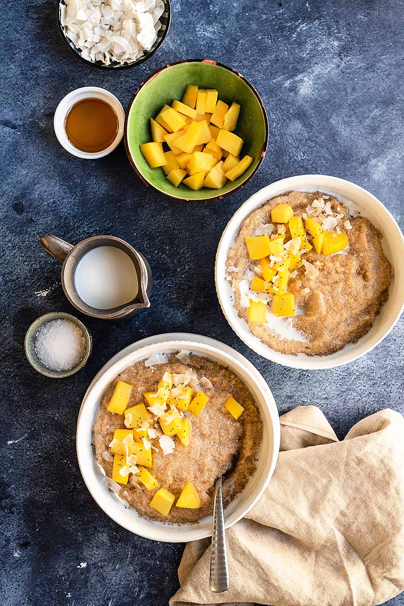 Vertical top-down image of two breakfast bowls with mango chunks and coconut shreds, next to bowls of milk, mango, and maple syrup.