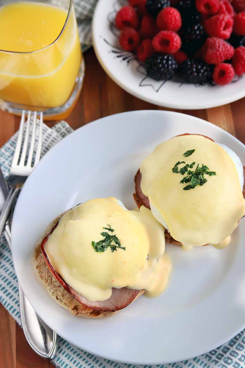 Vertical overhead image of two pieces of eggs Benedict on a white plate, with a cloth napkin and silverware to the left, and a glass of orange juice and a bowl of fruit at the top of the frame.