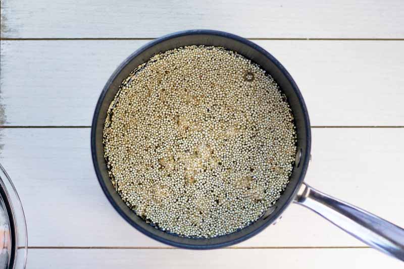 Overhead shot of uncooked quinoa in water, in a nonstick saucepan on a white painted wood surface.