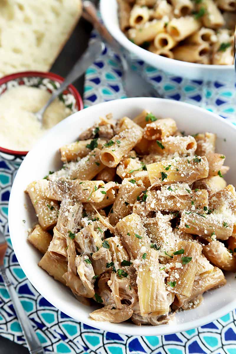 Vertical close-up image of a white bowl with cooked rigatoni and assorted garnishes on a blue patterned napkin next to a spoon in a bowl of grated cheese.