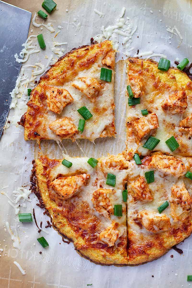Vertical top-down image of one slice of a cauliflower crust pizza topped with buffalo chicken chunks pulled away from the other three slices.