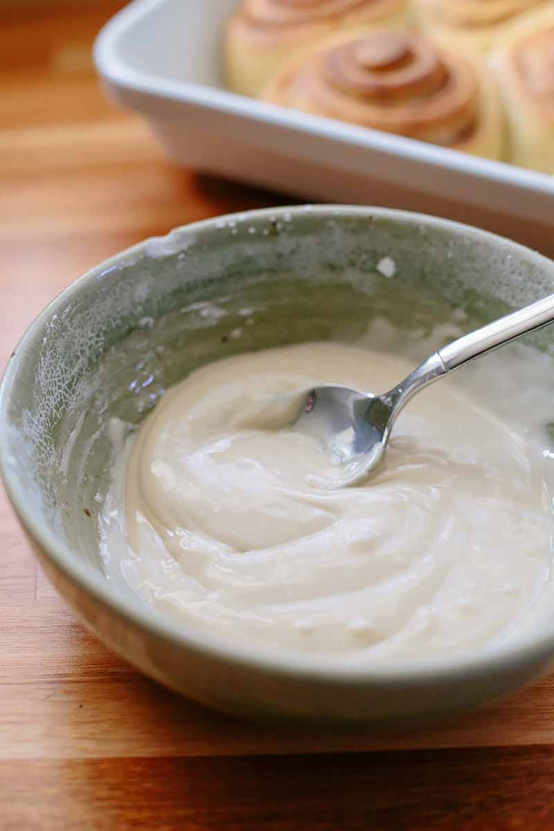 Vertical image of white icing and a spoon in a bowl on a wooden table.