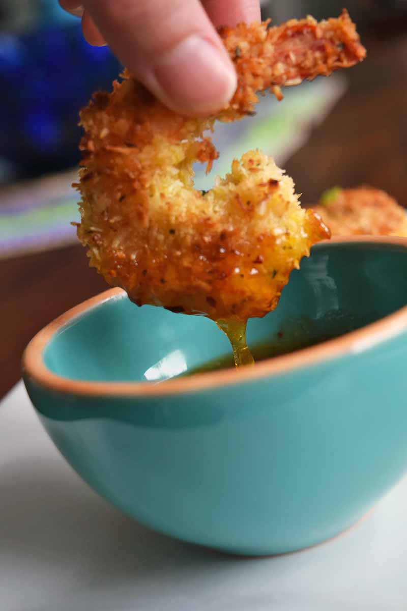 Vertical image of a hand dipping a panko coated baked shrimp into a blue bowl of sweet and sour sauce, on a white plate on a brown table.