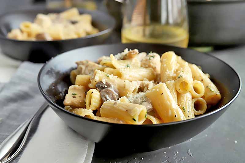 Horizontal image of a black bowls with creamy pasta, with a glass of white wine.