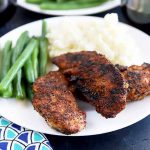 Horizontal image of a white plate with blackened tenders next to green beans and mashed potatoes.