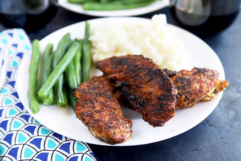 Horizontal image of a white plate with blackened tenders next to green beans and mashed potatoes.