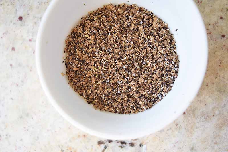Overhead horizontal image of a coffee and spice mixture in a white bowl, on a speckled beige countertop.