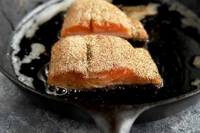 Horizontal image of two seasoned fish fillets cooking in a pan.