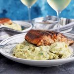 Horizontal image of a cooked salmon fillet on mashed potatoes in front of glasses of white wine.