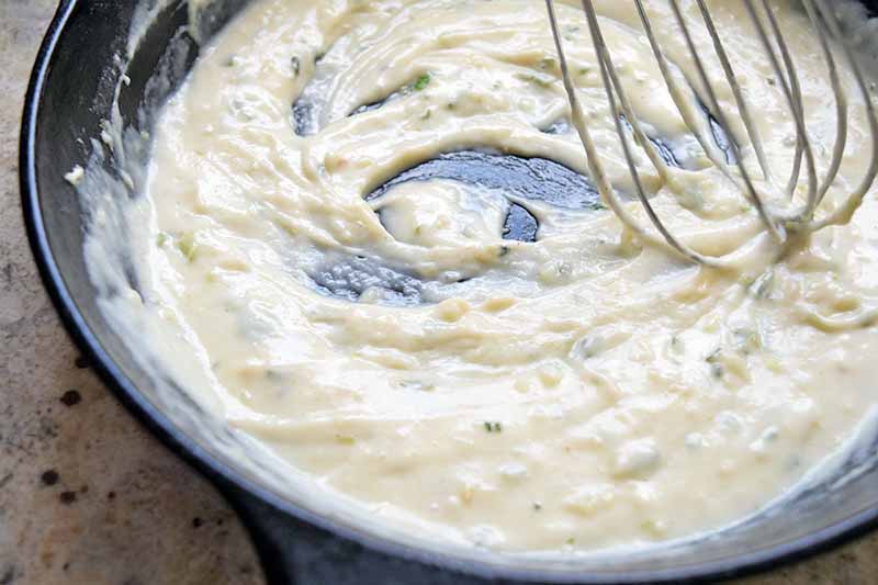 Horizontal image of a wire whisk stirring a creamy bechamel sauce in a large frying pan, on a beige speckled surface.