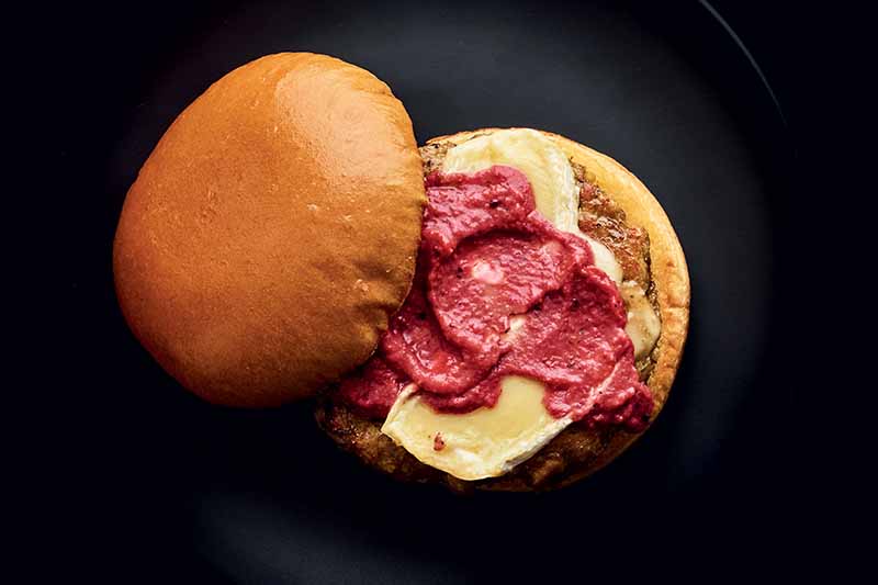 Overhead horizontal image of a chicken and mushroom burger with pink ketchup made with strawberries on top, on a burger bun, on a black plate with a black background.