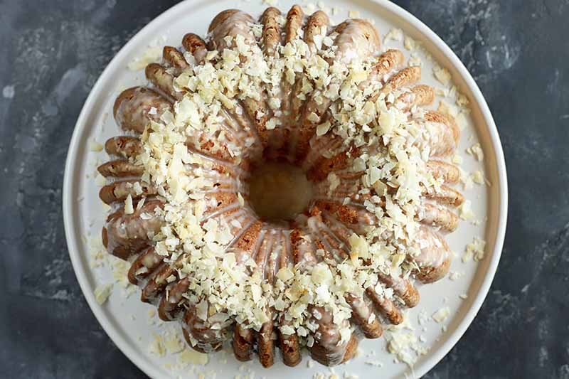 Horizontal image of a whole bundt cake with white glaze and white garnishes on a plate on a black surface.