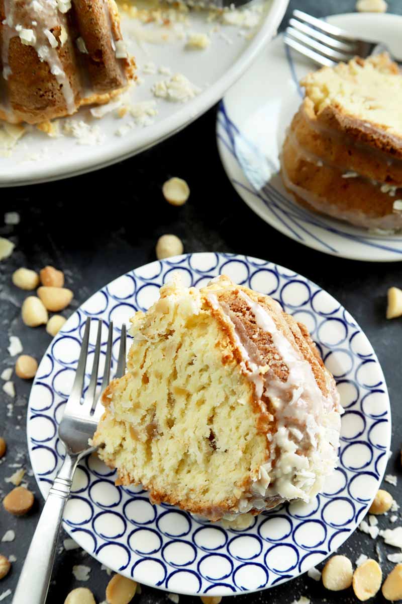 Vertical image of a slice of bundt cake with glaze on a patterned plate next to scattered nuts.
