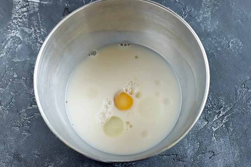 Non-dairy milk and an egg in a stainless steel mixing bowl, on a dark gray surface.
