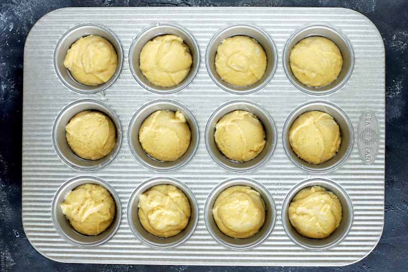 Cornbread batter portioned equally in each of twelve muffin wells in a metal tin, on a dark gray background.