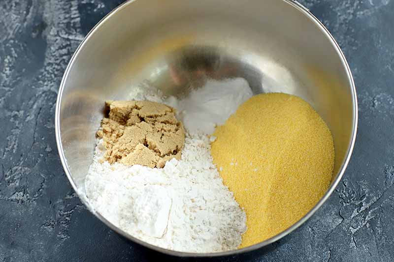 Yellow cornmeal, gluten-free flour, and light brown sugar in a large stainless steel mixing bowl, on a dark gray stone surface with marks and scratches.