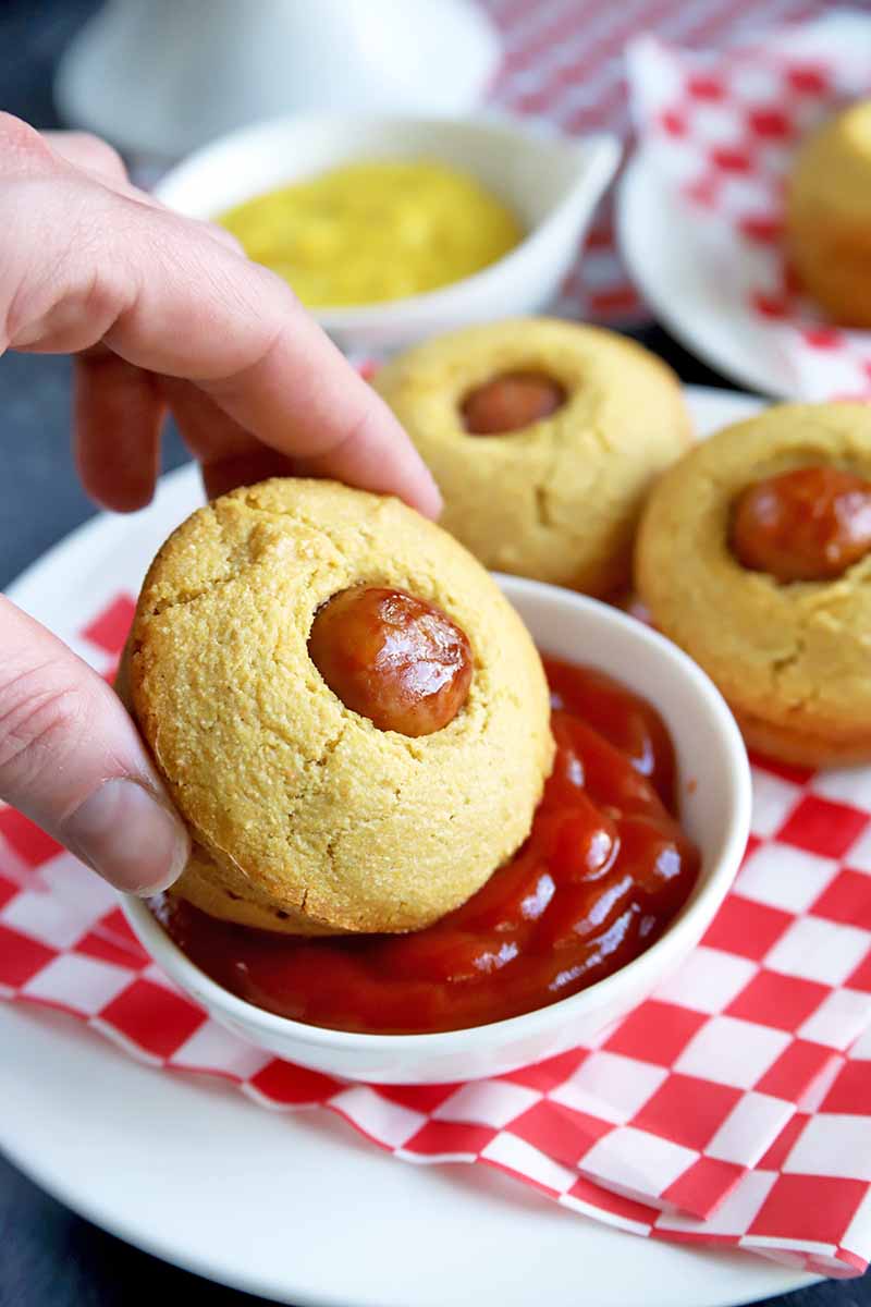 A hand dips a corn muffin with a hot dog in the center into a bowl of ketchup, with more muffins and a bowl of mustard in the background, on white plates topped with red and white checkered paper lines, on a gray surface.