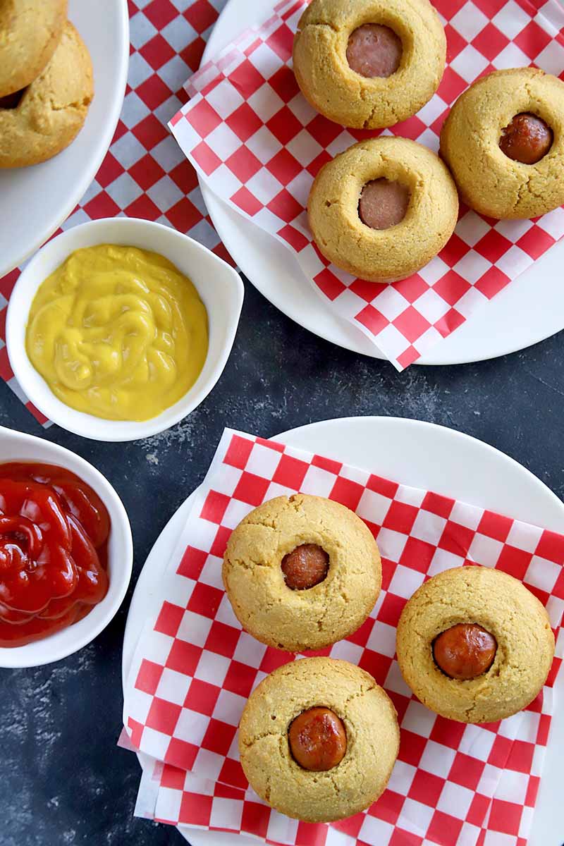 Overhead image of two white plates topped with red and white checkered paper liners and three corn dog muffins each, with another plate and two small white bowls of ketchup and mustard to the left, on a dark gray surface.