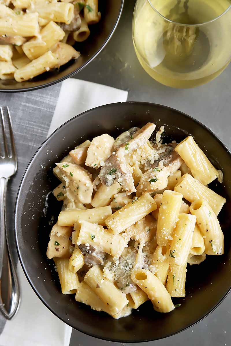 Vertical top-down image of black bowls with creamy rigatoni and mushrooms next to a fork and white napkin.