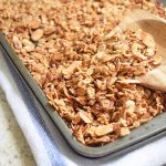 Horizontal image of a spoon on top of baked granola in a sheet pan resting on a white towel.
