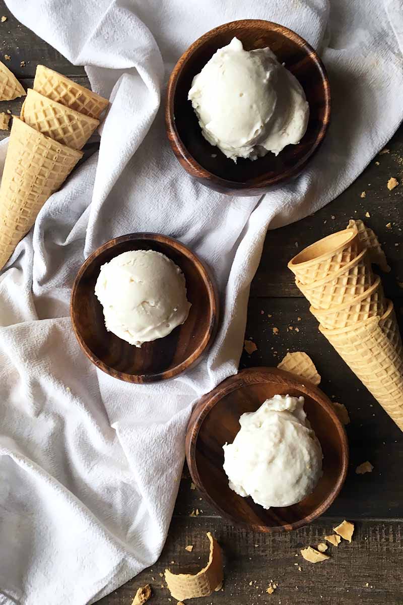 Vertical image of three wooden bowls with scoops of white ice cream on a white towel next to cones.