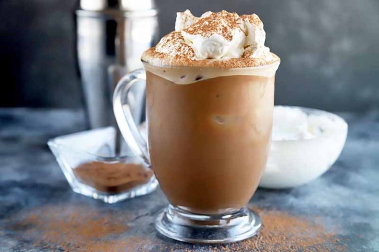 Horizontal image of a glass mocha drink topped with whipped cream and cocoa powder, next to a metal shaker, cocoa powder, and whipped cream.