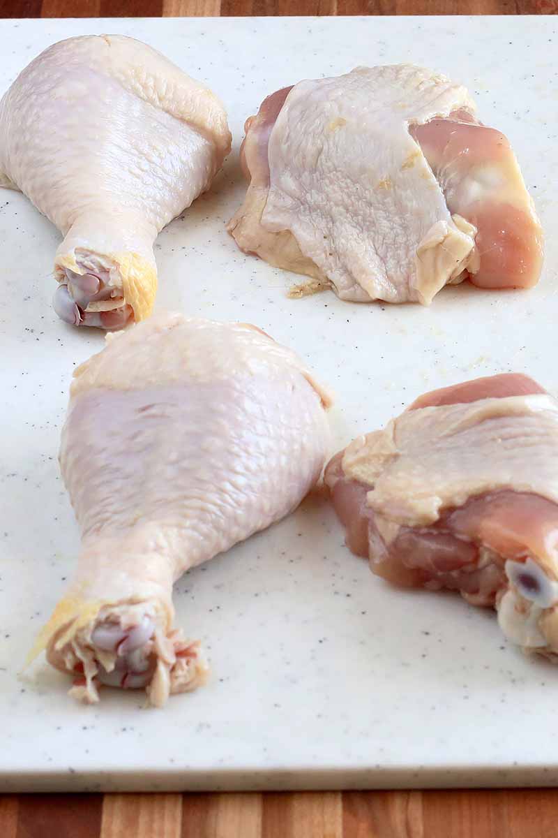 Vertical image of raw chicken drumsticks and thighs on a white cutting board.