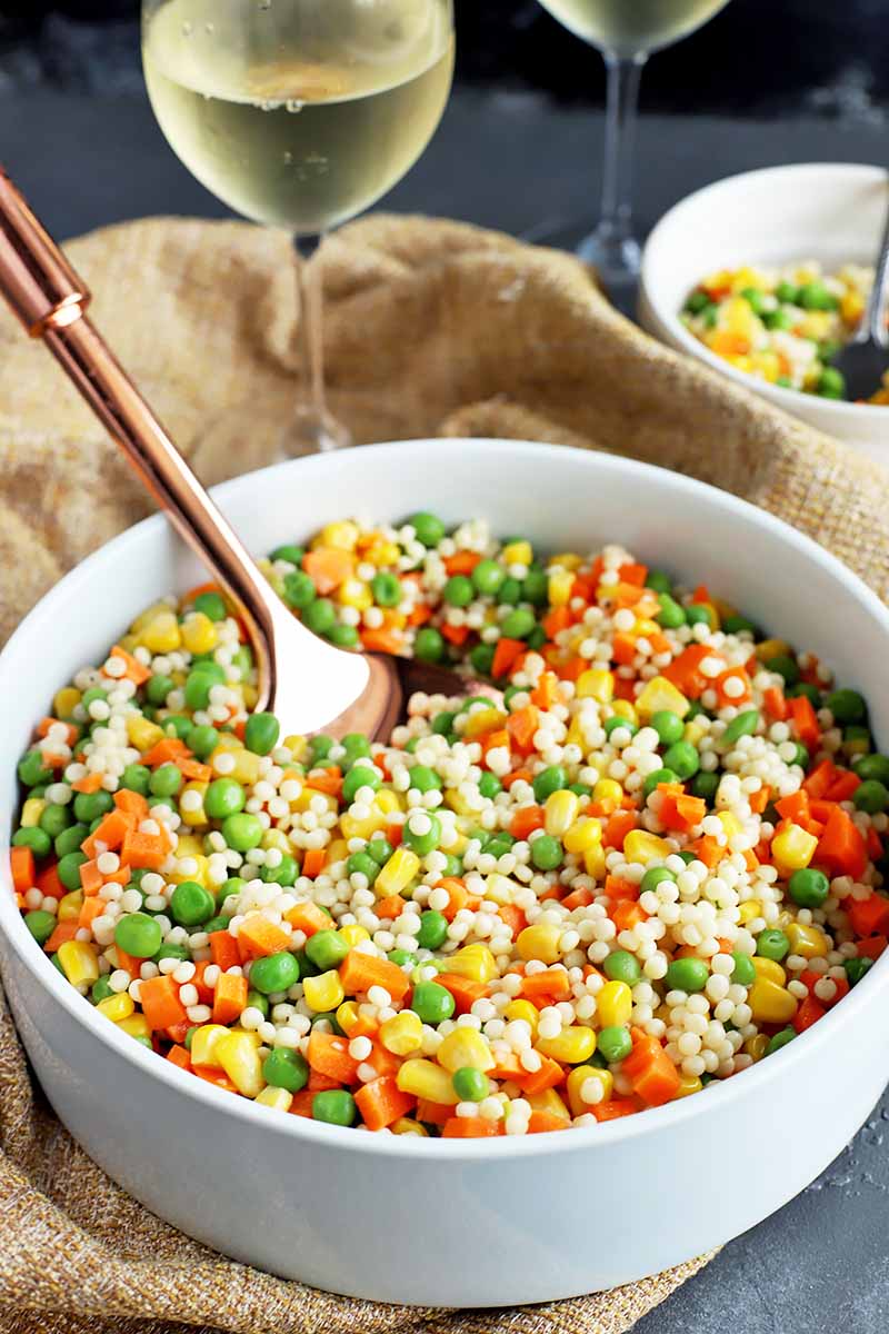 Vertical image of a large white bowl filled with a vegetable and grain medley with a spoon inserted into it, next to glasses of wine.