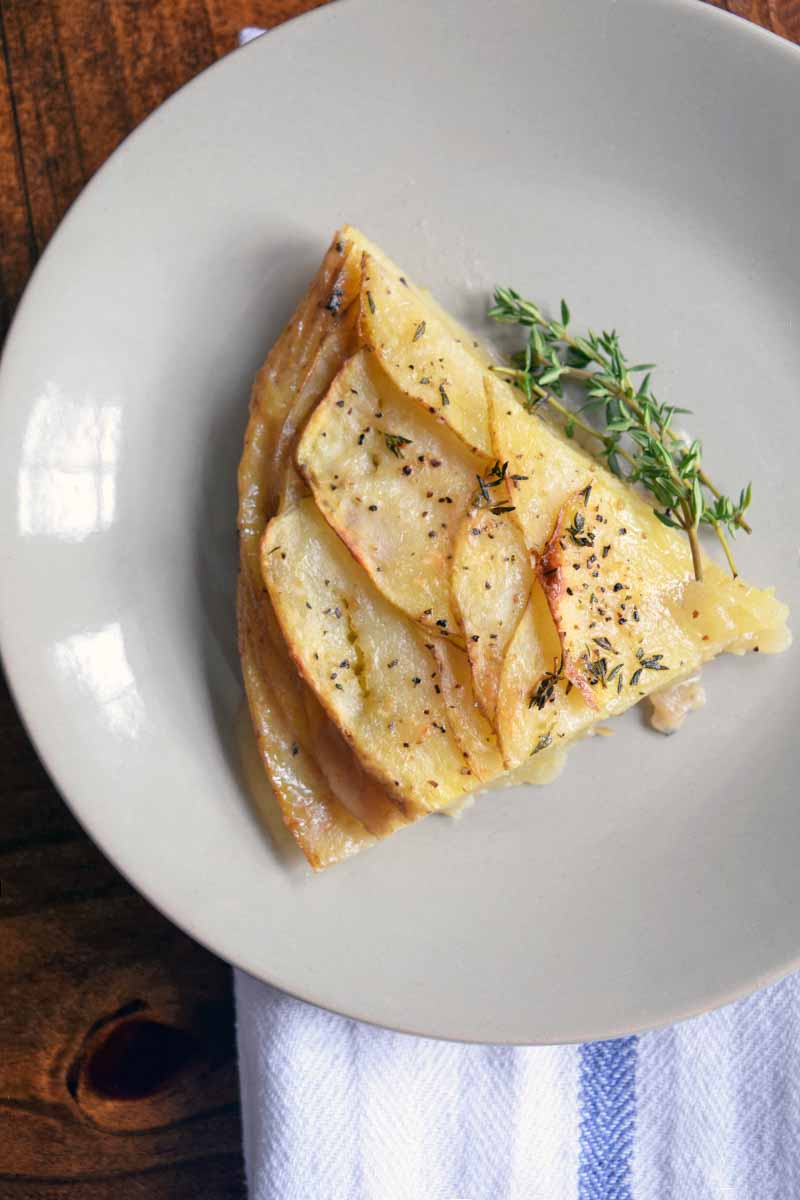 Overhead horizontal image of a triangular slice of potato tian with melted cheese on top and a sprig of fresh thyme, on a white plate, on top of a folded blue and white striped cloth on a brown wood table.
