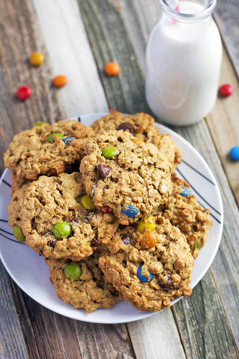 Oatmeal M&M cookies are piled on a white plate beside a glass bottle of milk, on a brown wood surface with scattered pieces of candy in various colors.