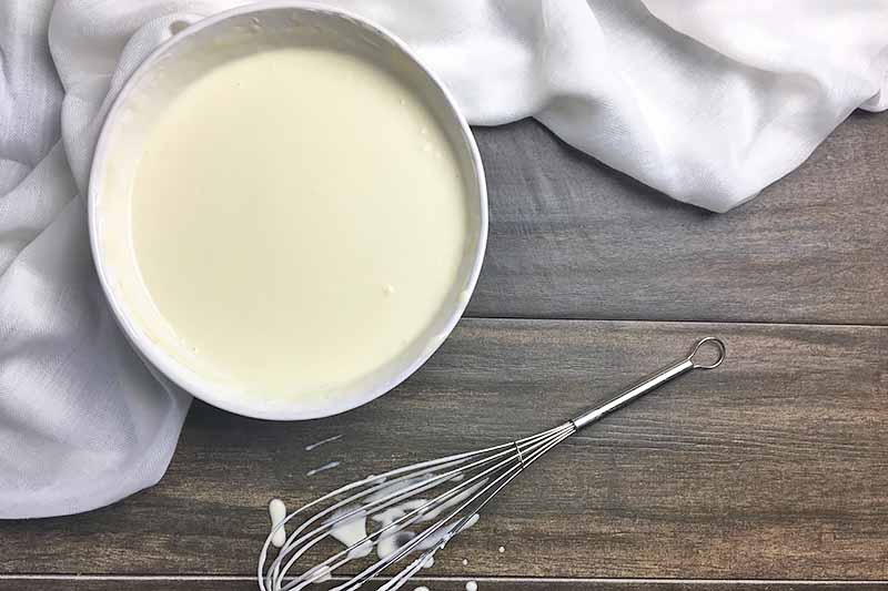 Horizontal image of a white bowl with a white liquid mixture next to a whisk with batter.