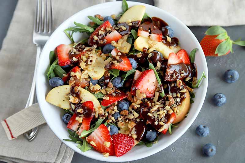 Overhead horizontal image of a shallow white bowl at the center of the frame, filled with arugula, fresh berries, apple slices, and chopped walnuts, with chocolate balsamic salad dressing on top, on a gray surface with two folded beige cloth napkins and a fork, with scattered fresh berries.