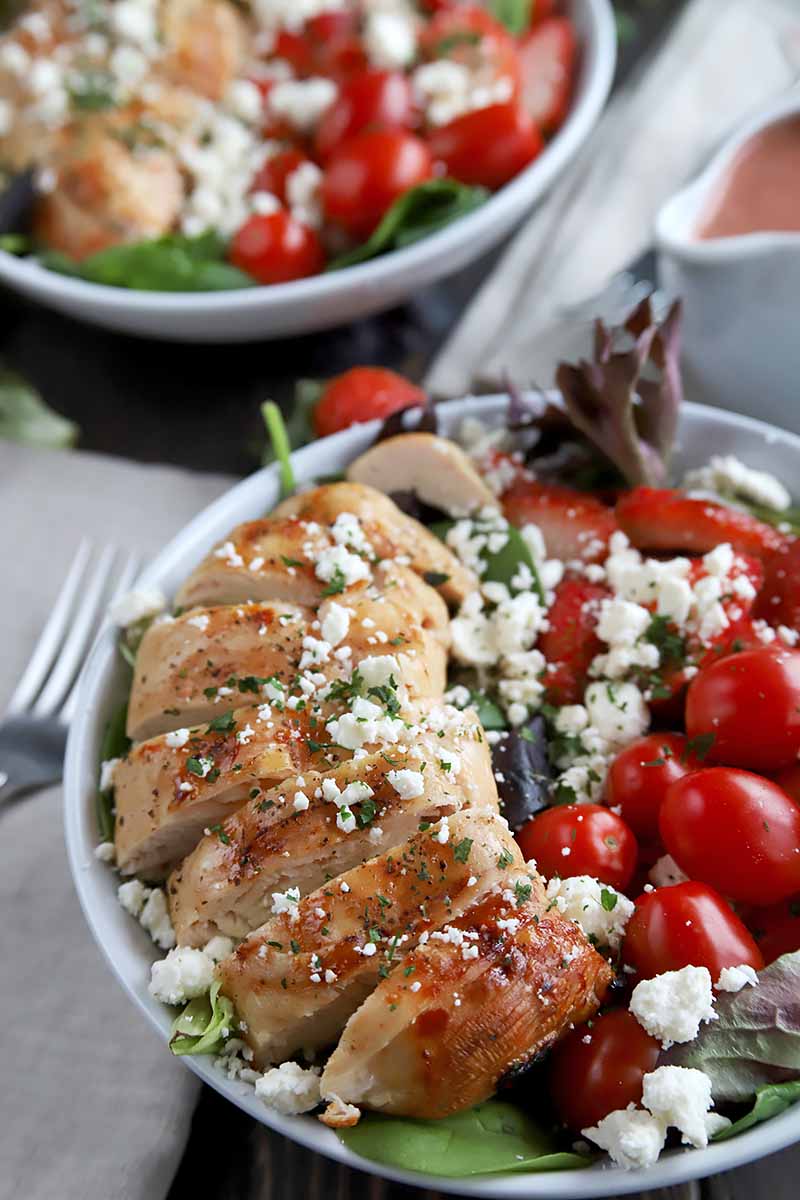Vertical image of two white plates with sliced chicken breasts topped with crumbled cheese next to sliced strawberries and grape tomatoes.