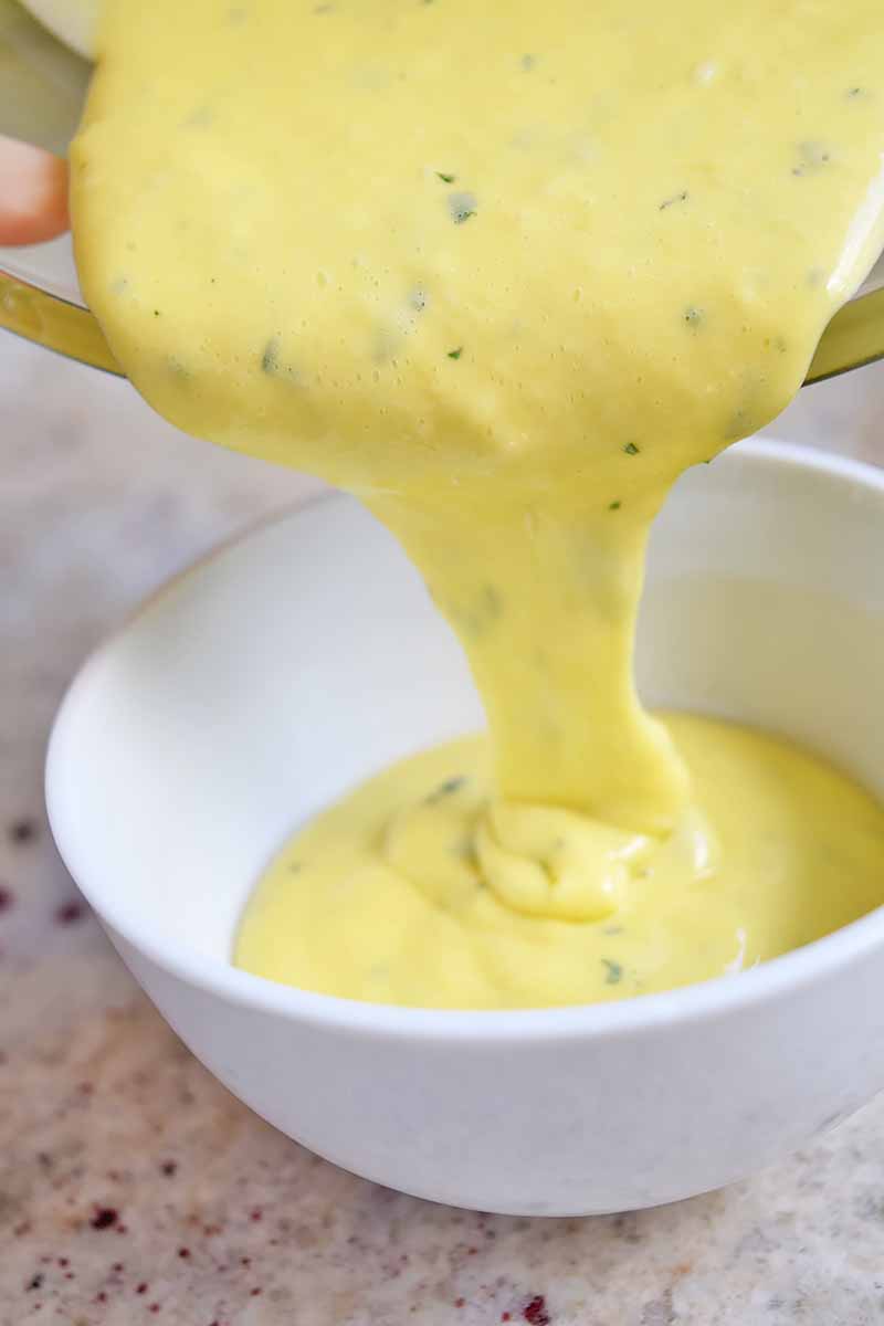 Creamy yellow aioli flecked with green chopped herbs being poured from a mixing bowl into a white serving dish, on a gray and brown speckled countertop.