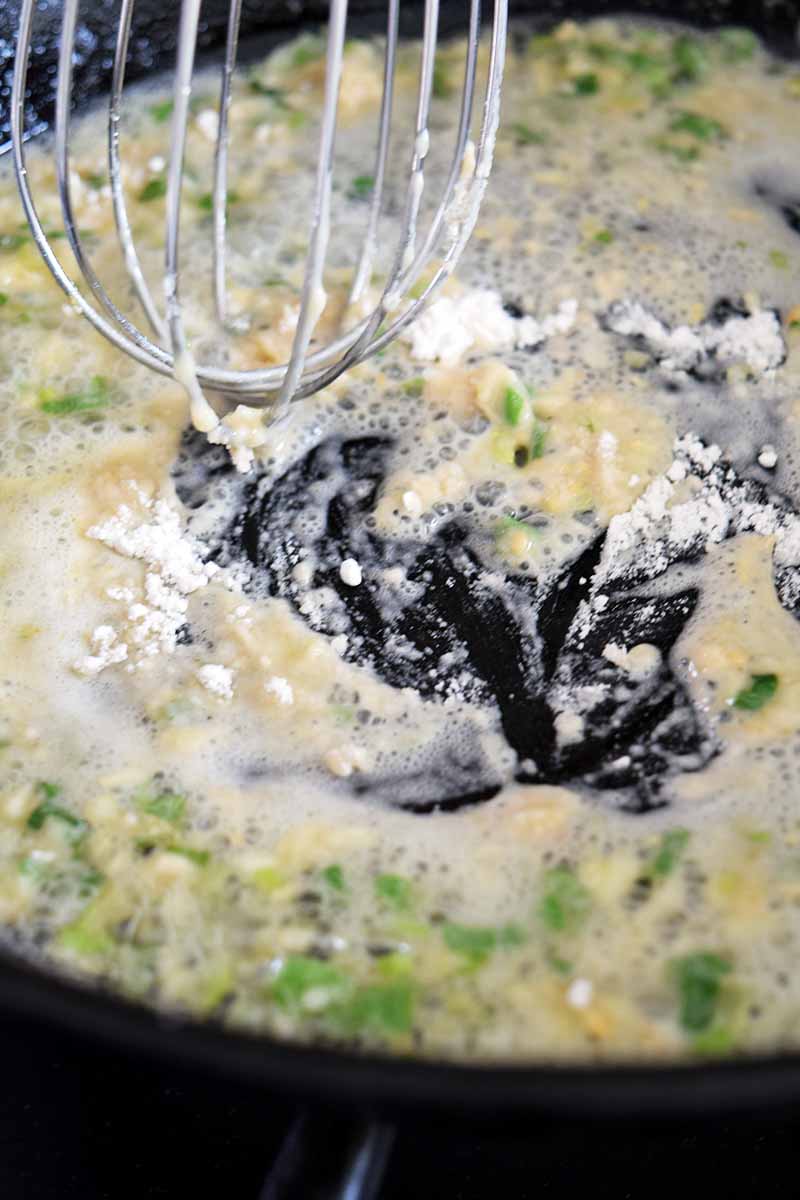 Vertical closeup image of a wire whisk stirring a mixture of melted butter, scallions, and flour in a large frying pan.
