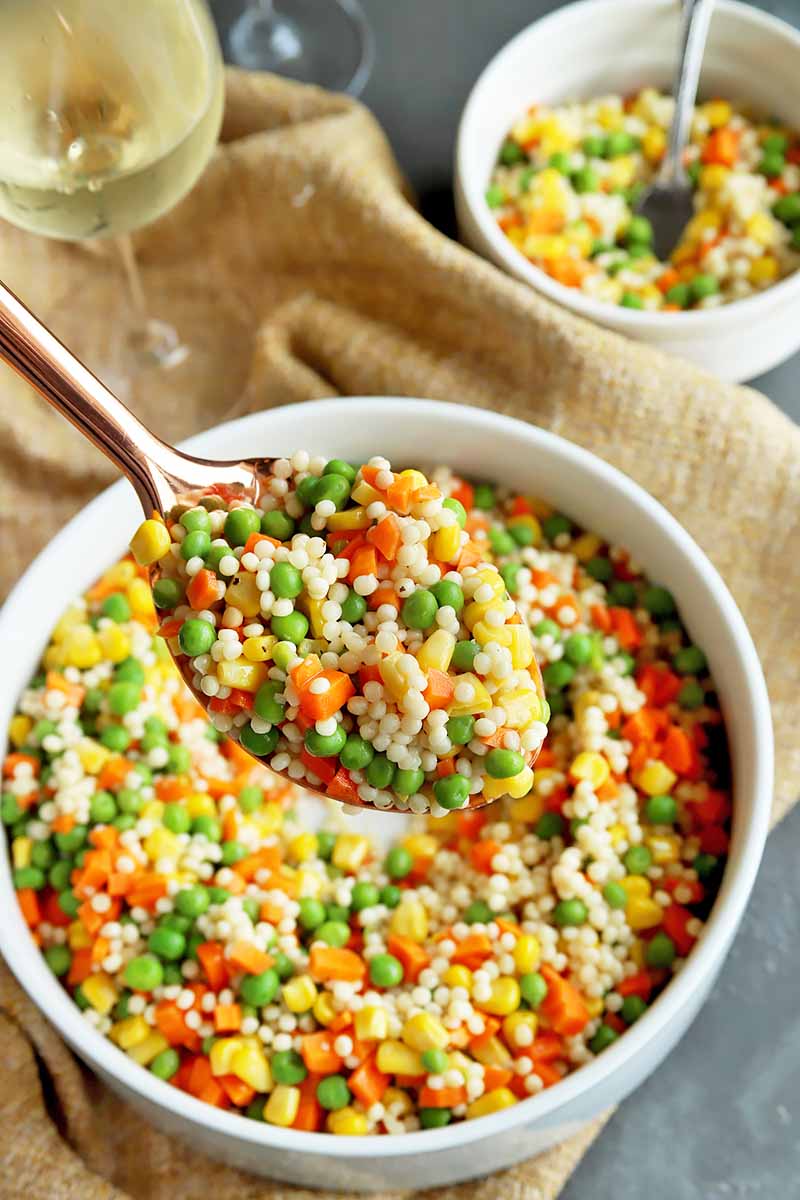 Vertical image of a bronze spoon holding a spoonful of a grain and veggie medley over a large bowl of the same dish.