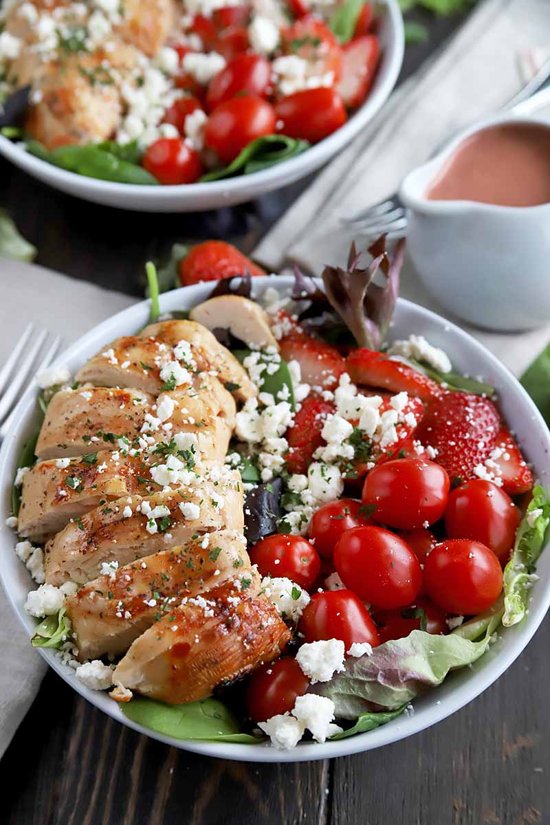 Vertical image of two plates with chicken salads with tomatoes and strawberries, next to white napkins and a spouted vessel with pink dressing.