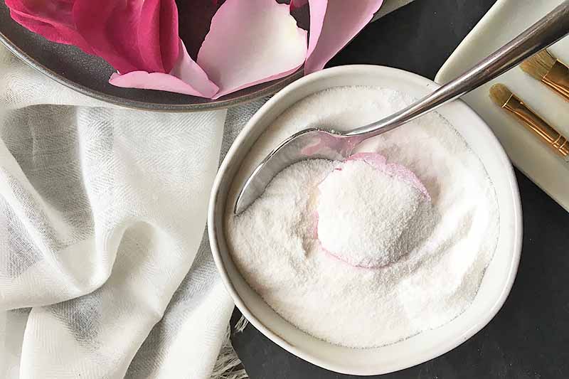 Horizontal image of a petal being covered in sugar by a metal spoon in a white bowl next to a white napkin and pink rose petals.