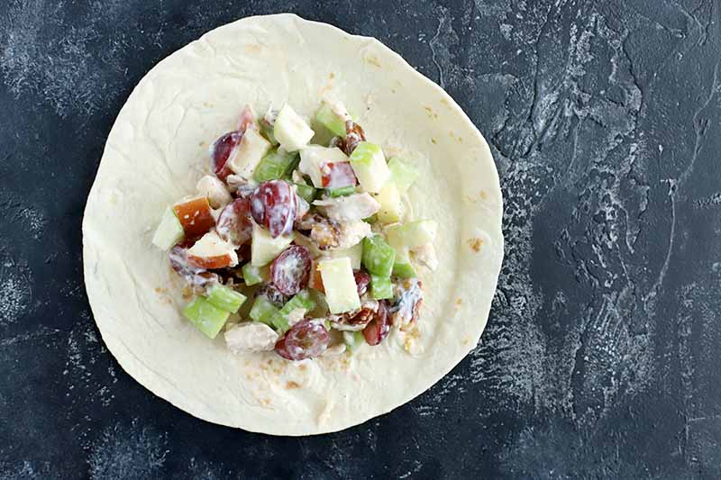 A large dollop of chicken salad with fruit and nuts is at the center of a flour tortilla, on a dark gray surface with white speckles and streaks.