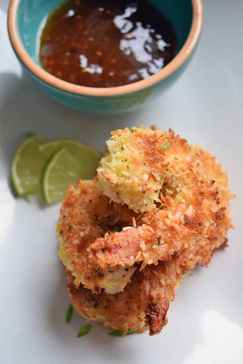 Vertical overhead image of a stack of three coconut shrimp, with slices of lime and a blue and terra cotta bowl of sweet and sour sauce on a white plate.