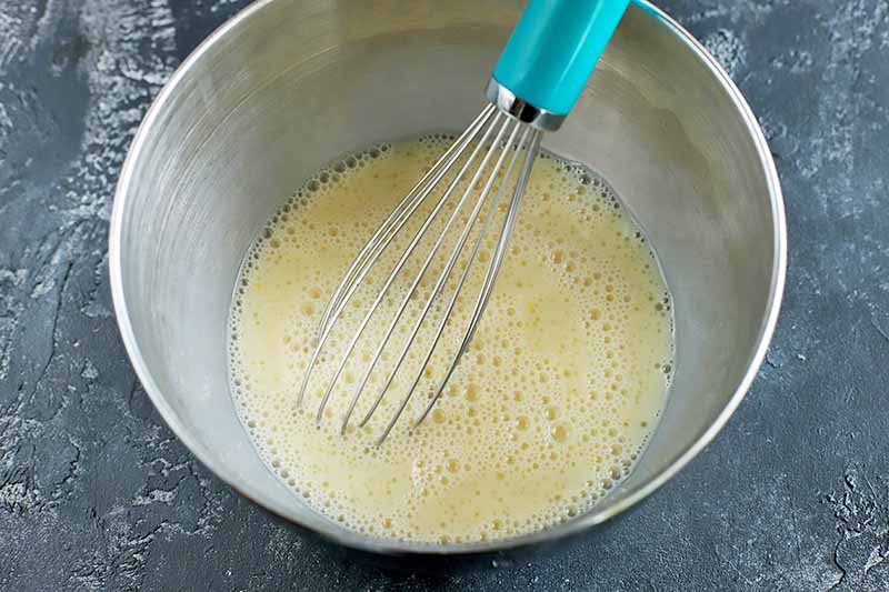Overhead closely cropped horizontal image of an egg and dairy-free milk mixture in a stainless steel mixing bowl, being stirred with a wire whisk with a blue handle, on a gray background.