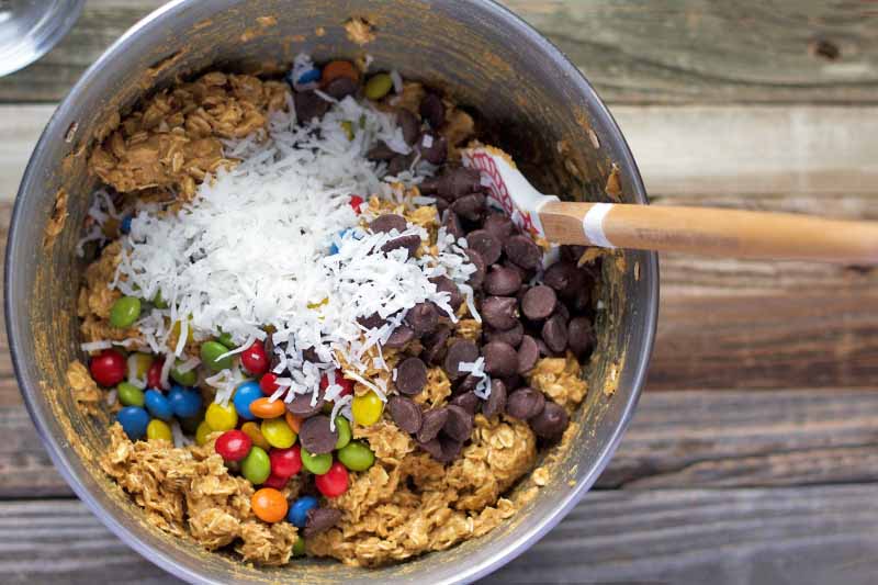 Horizontal closely cropped overhead image of a stainless steel bowl of an oat mixture with M&Ms, shredded coconut, and chocolate chips on top, being stirred with a spatula, on a wood surface.