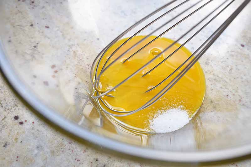 Closeup horizontal image of yellow egg and salt being mixed with a wire whisk in a bowl.